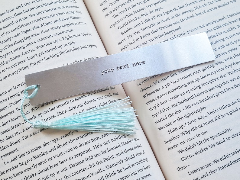 May include: A silver metal bookmark with a light blue tassel. The bookmark has the text "your text here" engraved on it.