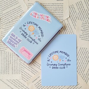 May include: A blue and white card with the text "Lifetime Member of the Grumpy Sunshine Book Club" in a whimsical font. The card features a cartoon sun and cloud with a lightning bolt and a heart.