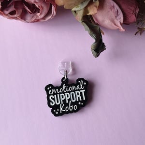 Könnte beinhalten: Ein schwarz-weißer Anhänger mit dem Text "emotional SUPPORT Kobo" in einer verspielten Schriftart. Der Anhänger ist an einem transparenten Kunststoffclip befestigt, vor einem hellvioletten Hintergrund mit floralen Akzenten.
