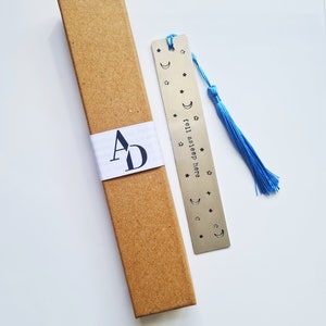May include: A silver bookmark with a blue tassel. The bookmark has a crescent moon and stars design and the text "fell asleep..." on it. The bookmark is in a brown cardboard box.