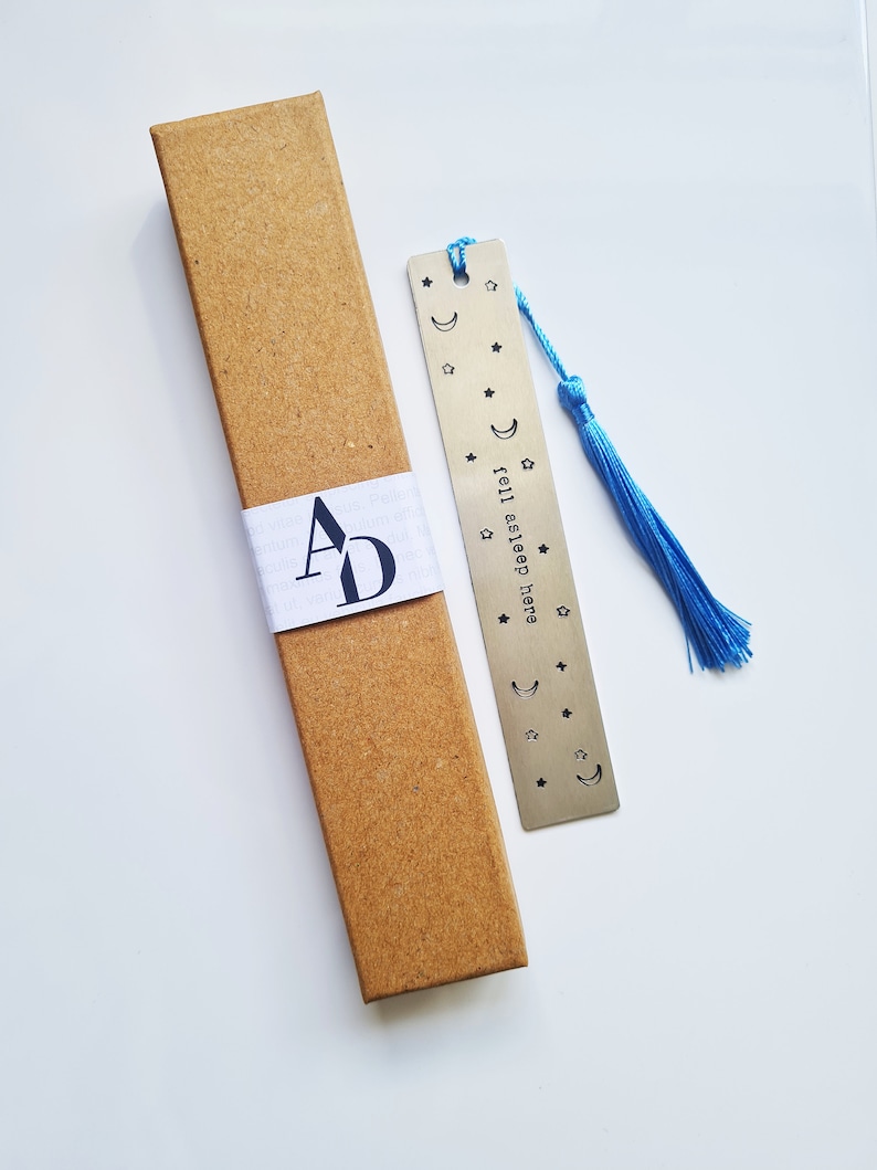 May include: A silver bookmark with a blue tassel. The bookmark has a crescent moon and stars design and the text "fell asleep..." on it. The bookmark is in a brown cardboard box.