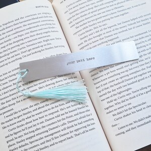 May include: A silver metal bookmark with a light blue tassel. The bookmark has the text "your text here" printed on it.