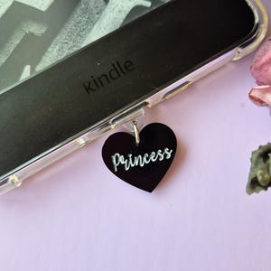 May include: A black Kindle e-reader with a clear case and a black heart-shaped charm that says "Princess" hanging from it.