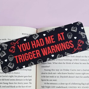 May include: Black and white bookmark with a red and white text overlay that reads "You Had Me At Trigger Warnings". The bookmark is decorated with hearts, skulls, roses, and guns.