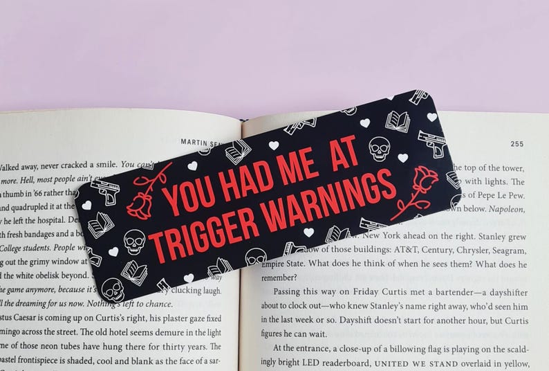 May include: Black and white bookmark with a red and white text overlay that reads "You Had Me At Trigger Warnings". The bookmark is decorated with hearts, skulls, roses, and guns.
