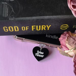 May include: A black heart-shaped charm with the text "Lotus Flower" in white script. The charm hangs from a clear phone case, with a book titled "God of Fury" in the background. Pink flowers are in the background.