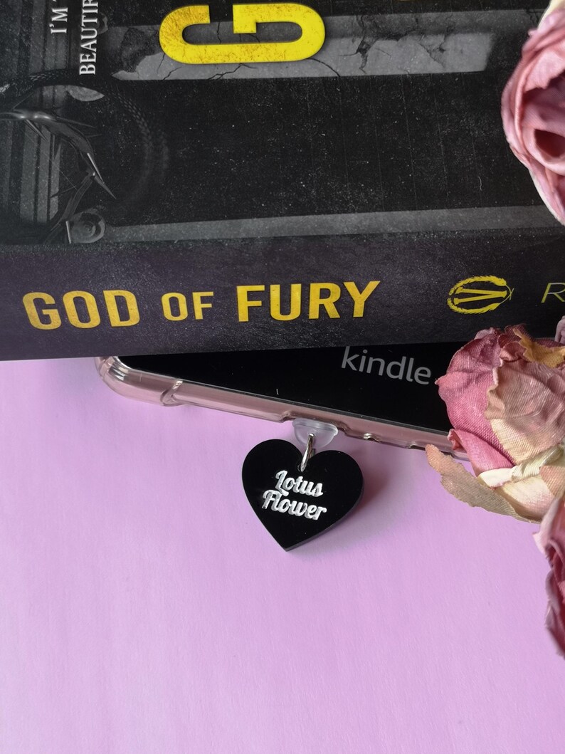 May include: A black heart-shaped charm with the text "Lotus Flower" in white script. The charm hangs from a clear phone case, with a book titled "God of Fury" in the background. Pink flowers are in the background.
