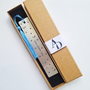 May include: A silver bookmark with a blue tassel and the text "fell asleep here" engraved on it. The bookmark is in a brown cardboard box with a white label that says "AD".