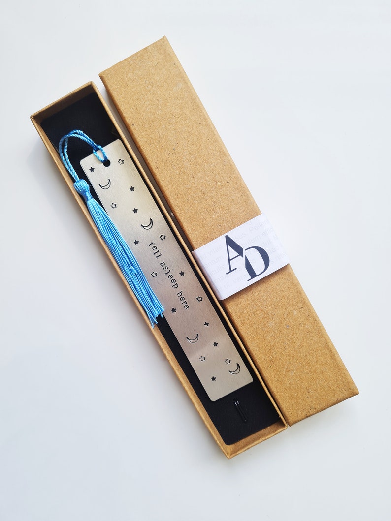 May include: A silver bookmark with a blue tassel and the text "fell asleep here" engraved on it. The bookmark is in a brown cardboard box with a white label that says "AD".