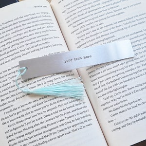 May include: A silver metal bookmark with a light blue tassel. The bookmark has the text "your text here" printed on it.