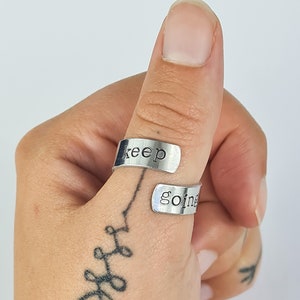 May include: A silver thumb ring with the words "keep going" engraved on it. The ring is worn on a person's thumb.