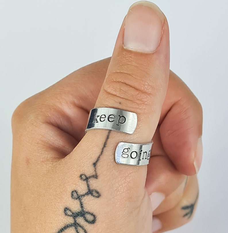 May include: A silver thumb ring with the words "keep going" engraved on it. The ring is worn on a person's thumb.