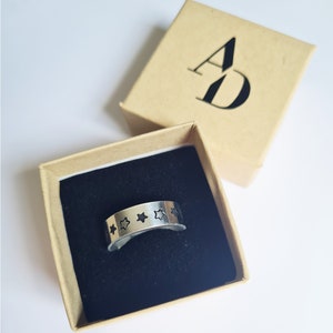 May include: A silver ring with five black star cutouts in a brown box with the letters "AD" printed on the top.