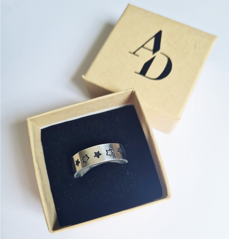 May include: A silver ring with five black star cutouts in a brown box with the letters "AD" printed on the top.