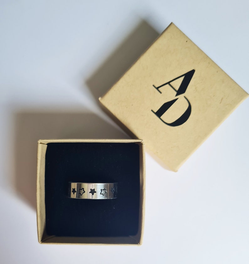 May include: A silver ring with a black star design on a black velvet background in a brown box with the letters "AD" printed on the top.