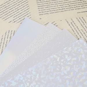 Puede incluir: Primer plano de tres papeles blancos diferentes con varios patrones. El papel de abajo tiene un patr&oacute;n hologr&aacute;fico de peque&ntilde;as formas iridiscentes. El papel del medio tiene un patr&oacute;n sutil y moteado. El papel de arriba es blanco liso.