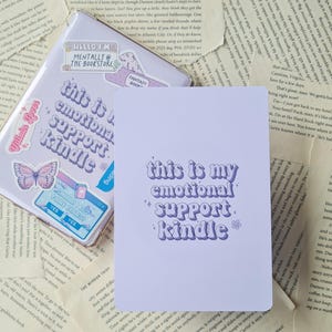 Puede incluir: Cuaderno morado y blanco con el texto "This is my emotional support kindle" en una fuente retro. El cuaderno tiene una cubierta de color morado claro y est&aacute; decorado con pegatinas.