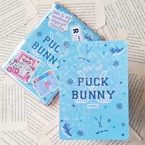 May include: A blue and white sticker with the text "Puck Bunny" in a white outline. The sticker has a hockey stick design and snowflakes. The sticker is on a blue background with a glittery effect.