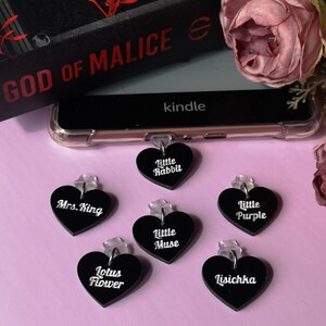 May include: A collection of black heart-shaped charms with white text, including "Little Rabbit," "Mrs. King," and "Lotus Flower." A Kindle device and a book titled "GOD OF MALICE" are in the background, along with a pink flower.