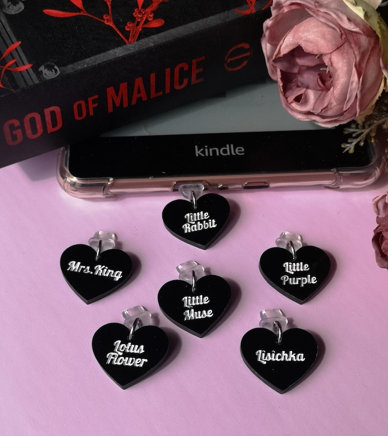 May include: A collection of black heart-shaped charms with white text, including "Little Rabbit," "Mrs. King," and "Lotus Flower." A Kindle device and a book titled "GOD OF MALICE" are in the background, along with a pink flower.