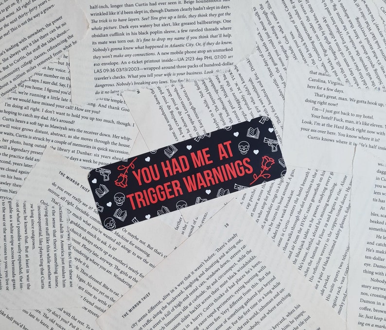 May include: A black and red bookmark with the text "You Had Me At Trigger Warnings" in white letters. The bookmark is decorated with skulls, guns, and a rose.
