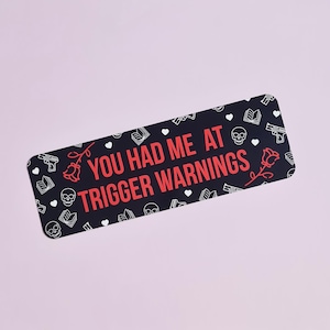 May include: Black and white sticker with a red text overlay that reads "You had me at trigger warnings". The sticker is decorated with a repeating pattern of hearts, roses, skulls, books, and guns.