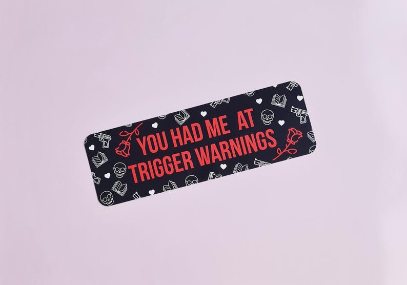 May include: Black and white sticker with a red text overlay that reads "You had me at trigger warnings". The sticker is decorated with a repeating pattern of hearts, roses, skulls, books, and guns.
