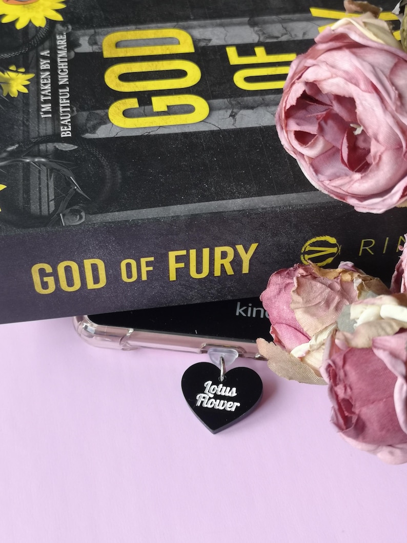 May include: A black heart-shaped charm with the words "Lotus Flower" in white script hangs near a book titled "God of Fury." The book has gold lettering and is partially obscured. Pink roses and a phone case are also visible.