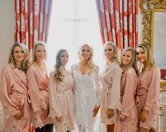 May include: Seven people, including a bride, stand before a window. The bride wears a white lace dress, while the others wear matching blush-coloured robes with lace trim. The setting appears to be a wedding.