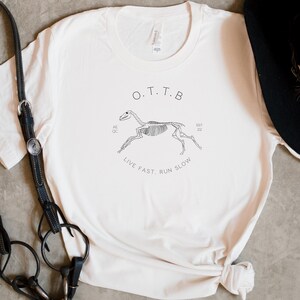May include: Off-white t-shirt with a black graphic of a running horse skeleton. The text "O.T.T.B" is above the graphic, with "LIVE FAST, RUN SLOW" below. A black hat and equestrian equipment are visible.