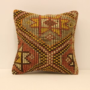 May include: Square decorative pillow with a woven geometric pattern in shades of brown, red, yellow, and gray. The pillow has a textured appearance and a symmetrical design, suitable for home decor.