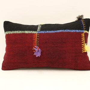 May include: A burgundy rectangular pillow with black, green, blue, and yellow embroidered details. The pillow has a fringe detail on the corners.