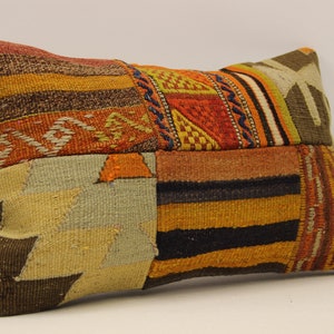 Op de afbeelding: Rechthoekig patchwork kussen met een verscheidenheid aan kleuren en patronen. Het kussen heeft een mix van oranje, bruine, gele en beige tinten, met geometrische en gestreepte ontwerpen. Het kussen is waarschijnlijk gemaakt van geweven stof.