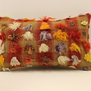 May include: A rectangular pillow with a red and brown checkered pattern. The pillow is decorated with colorful fluffy tassels in shades of red, orange, yellow, white, purple, and brown.