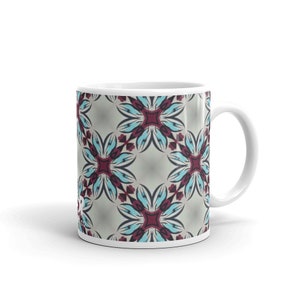 Puede incluir: Taza de cerámica blanca con un patrón geométrico en tonos azules, grises y burdeos. El patrón presenta un diseño repetitivo de flores estilizadas.