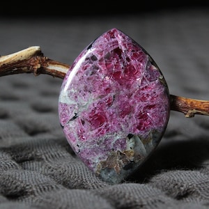 May include: A polished gemstone cabochon in shades of pink and magenta. The stone features a distinctive, irregular pattern with grey and black inclusions. The cabochon is displayed against a neutral backdrop, accentuating its natural beauty.