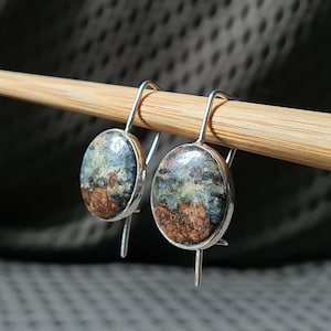 Può includere: Un paio di orecchini pendenti in argento con un design unico e astratto. Gli orecchini presentano una pietra rotonda blu, verde e marrone con una superficie testurizzata.