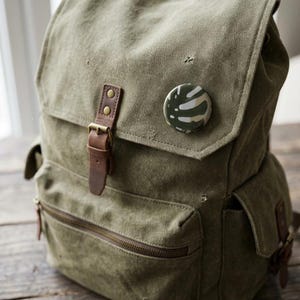 May include: A green canvas backpack with a flap closure, brown leather straps, and a front zipper pocket. A round button with a camouflage pattern is attached to the flap. The backpack has a vintage, worn look.