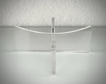 May include: A clear acrylic display stand with a vertical support and a curved top. The stand is designed to hold items and is made of transparent material. The stand is on a white surface.