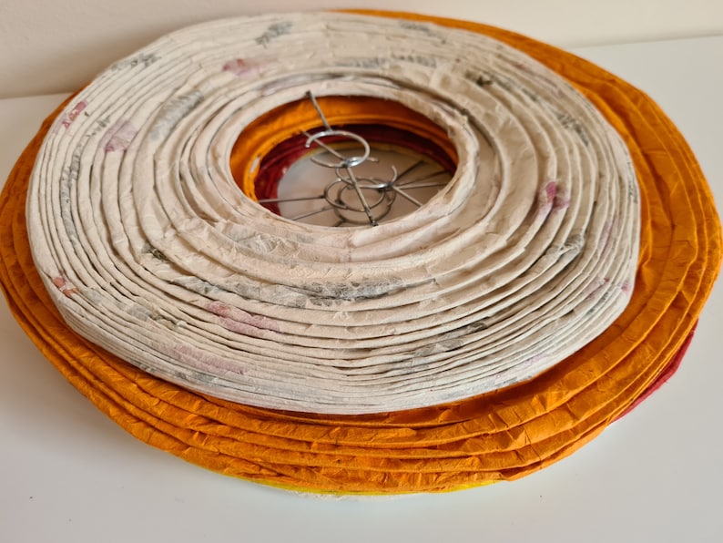 May include: A stack of paper lanterns in shades of orange, cream, and red. The cream-colored lantern has a floral pattern. The lanterns are circular and have a metal wire frame. The lanterns are stacked on top of each other.
