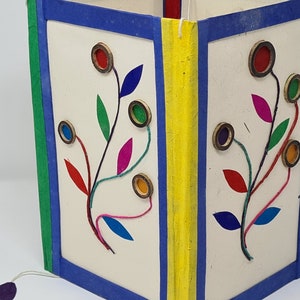 May include: A handmade journal with a white cover and colorful floral designs. The journal has a blue and green border and a yellow spine.