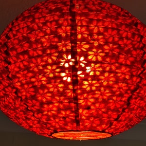 May include: A round, red paper lantern with a floral pattern. The lantern is lit from within, creating a warm, inviting glow. The design is visible through the translucent paper, adding a decorative element.
