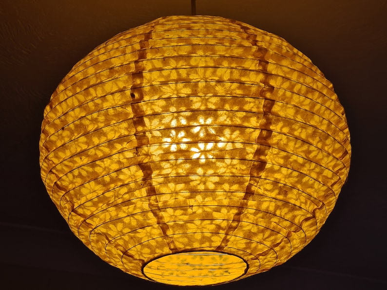 May include: A round, illuminated paper lantern with a warm yellow glow. The lantern's surface is patterned with floral cutouts, creating a soft, ambient light. The lantern is suspended from the ceiling, casting a gentle light.