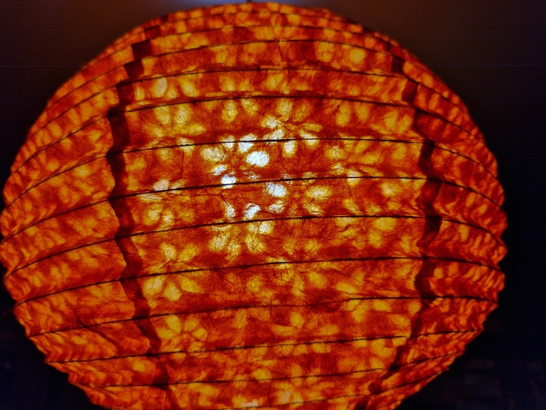 May include: A spherical, orange paper lantern with a ribbed design. The lantern's surface has a textured appearance, with visible patterns and a warm, inviting glow. The light source is visible through the paper, creating a soft, ambient light.