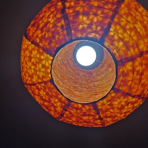 May include: A low-angle view of a round, orange paper lantern with a light bulb visible inside. The lantern has a patterned design and is suspended against a dark background, creating a warm, inviting ambiance. The light bulb is bright.