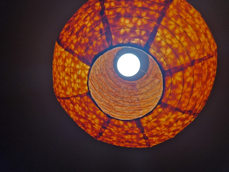 May include: A low-angle view of a round, orange paper lantern with a light bulb visible inside. The lantern has a patterned design and is suspended against a dark background, creating a warm, inviting ambiance. The light bulb is bright.