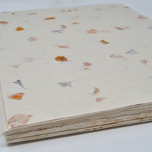 May include: A stack of handmade paper with a cream background and scattered orange and purple flower petals.