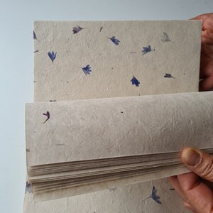 May include: A stack of handmade paper with a textured surface and embedded dried flowers. The paper is beige with blue and purple flower petals.