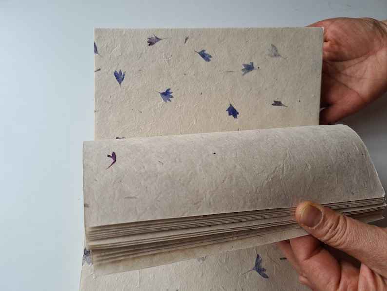May include: A stack of handmade paper with a textured surface and embedded dried flowers. The paper is beige with blue and purple flower petals.