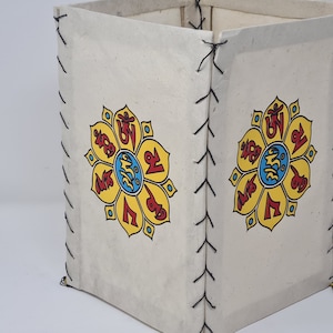 May include: A square, white fabric box with black stitching. The box has a yellow flower design with red and blue symbols on each side. The box has a black tassel.